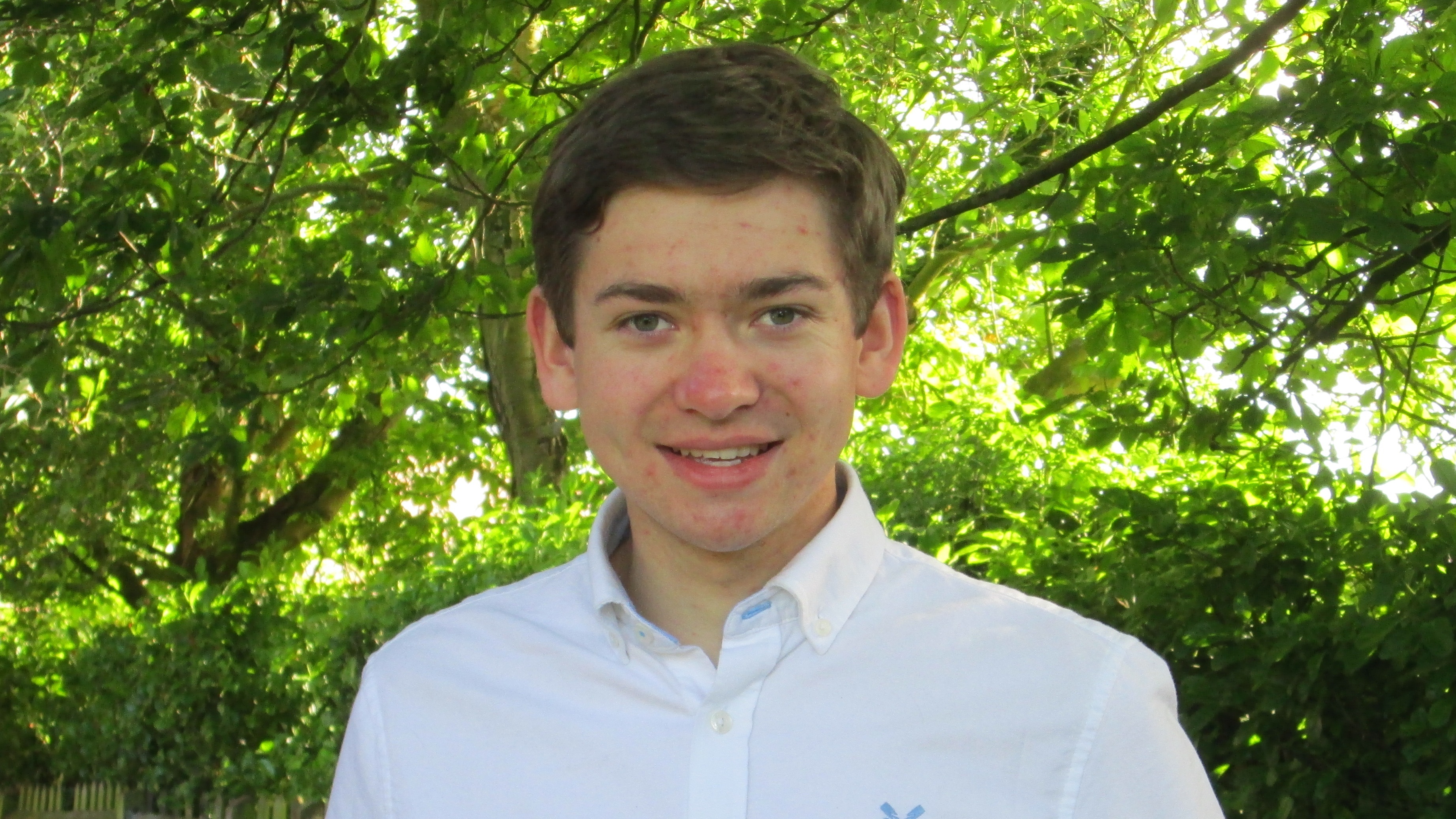 Head and shoulders image of young male student facing forward and smiling against a backdrop of green hedges