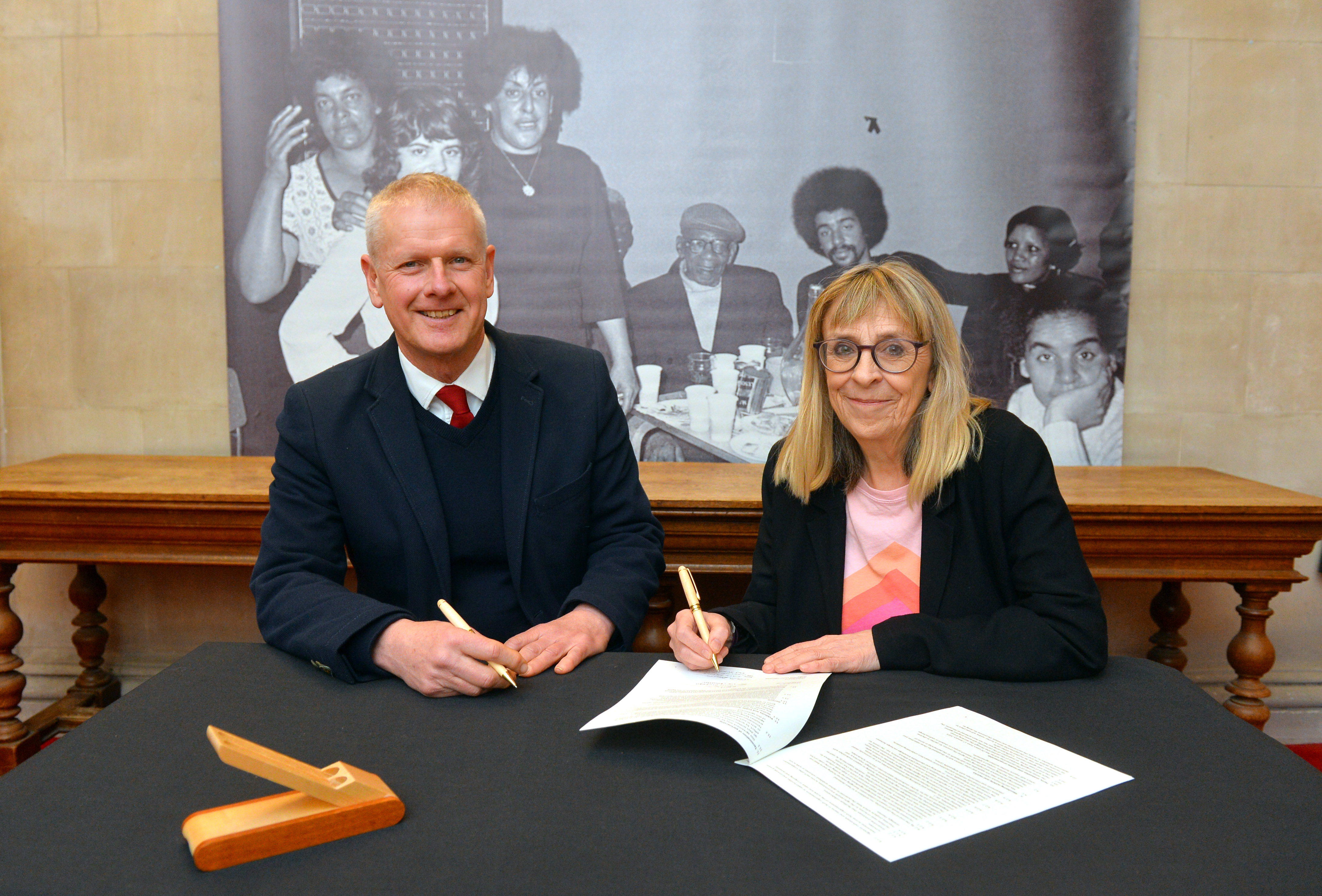 A man and a woman look at the camera as they sign a document on the table in front of them