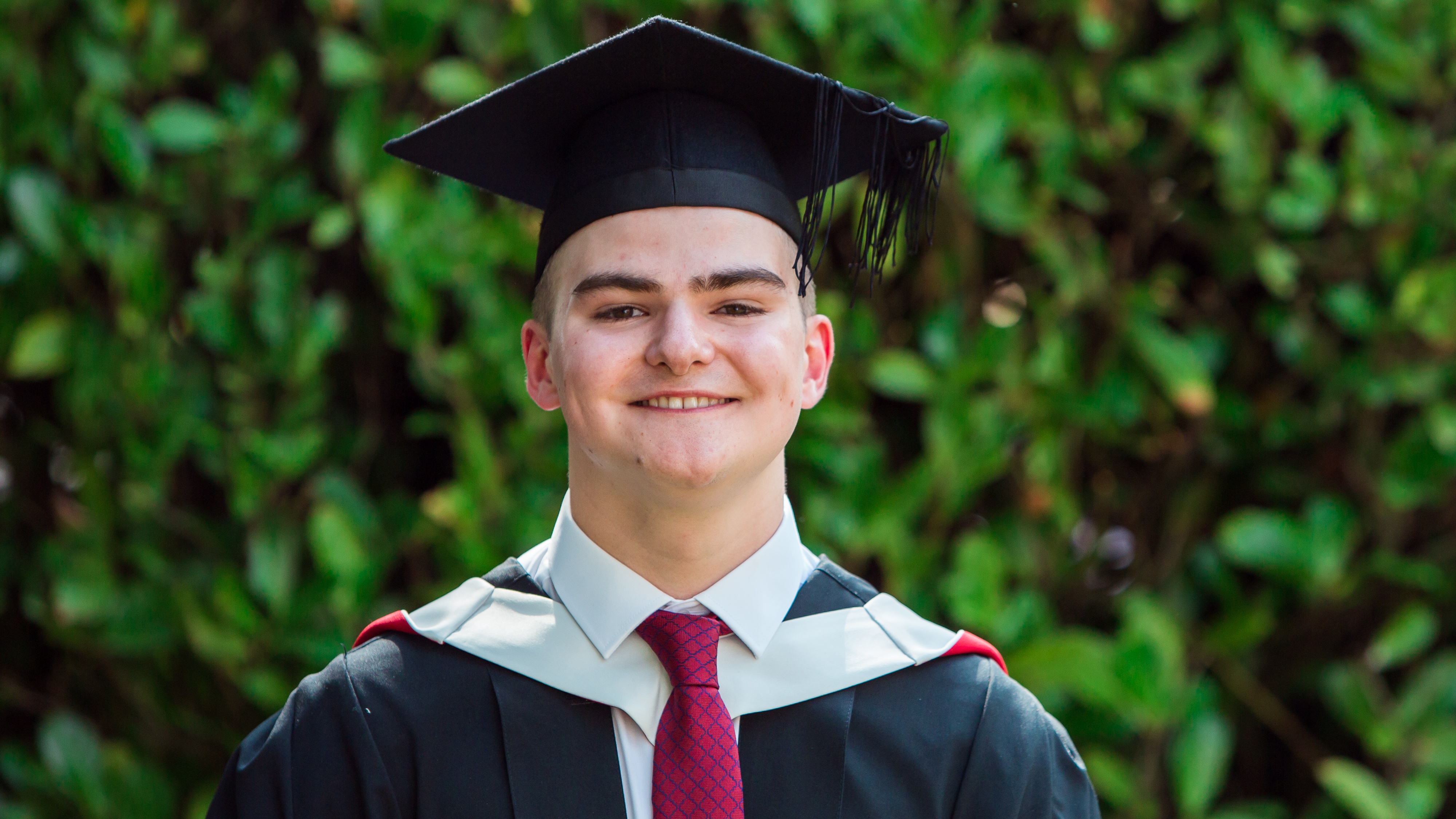 Young male graduate stood wearing graduation cap and gown in front of a green hedge