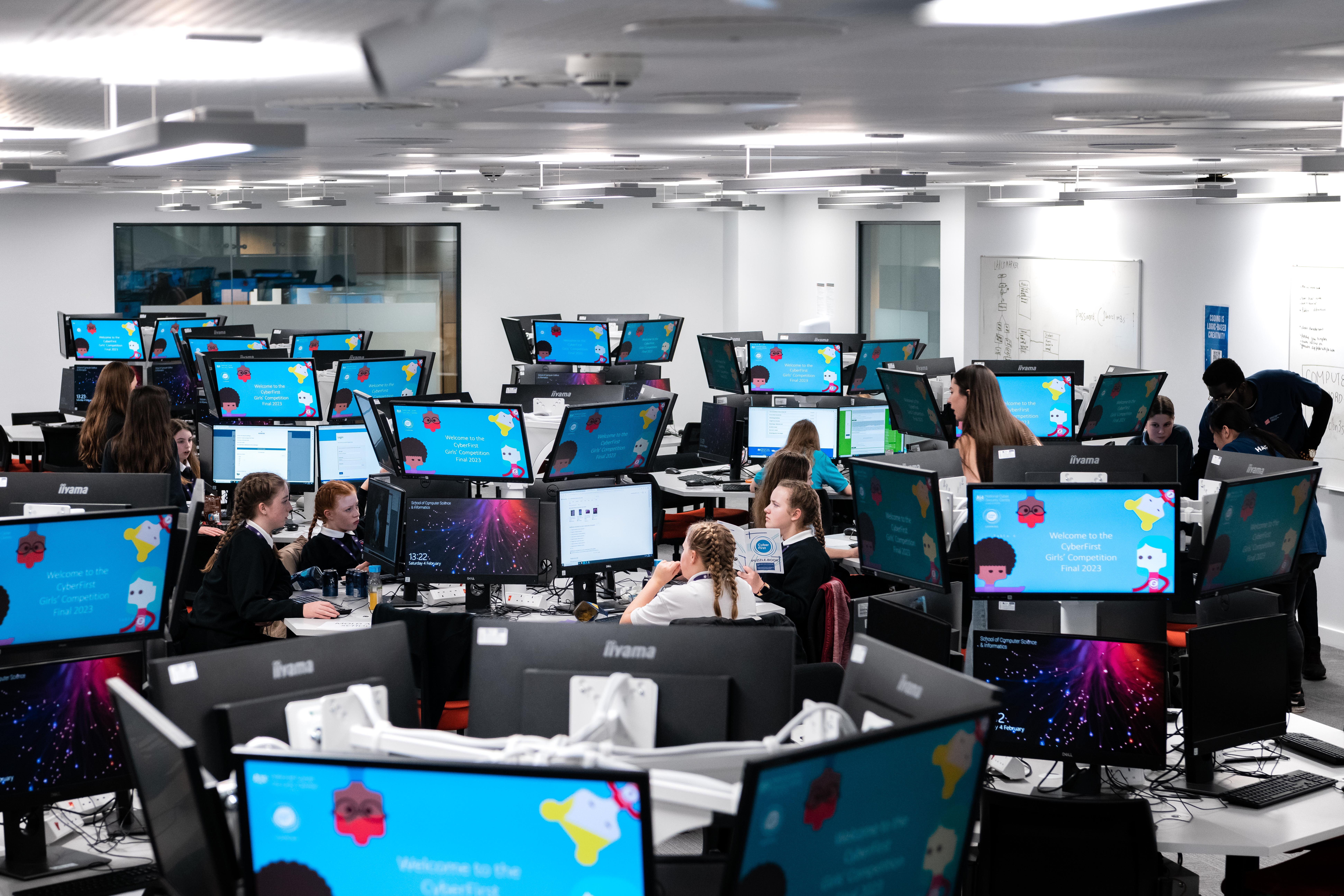 Image of mounted computer displays featuring the National Cyber Security Centre logo, cartoon characters and a message, which reads: Welcome to the CyberFirst Girls&rsquo; Competition Final 2023