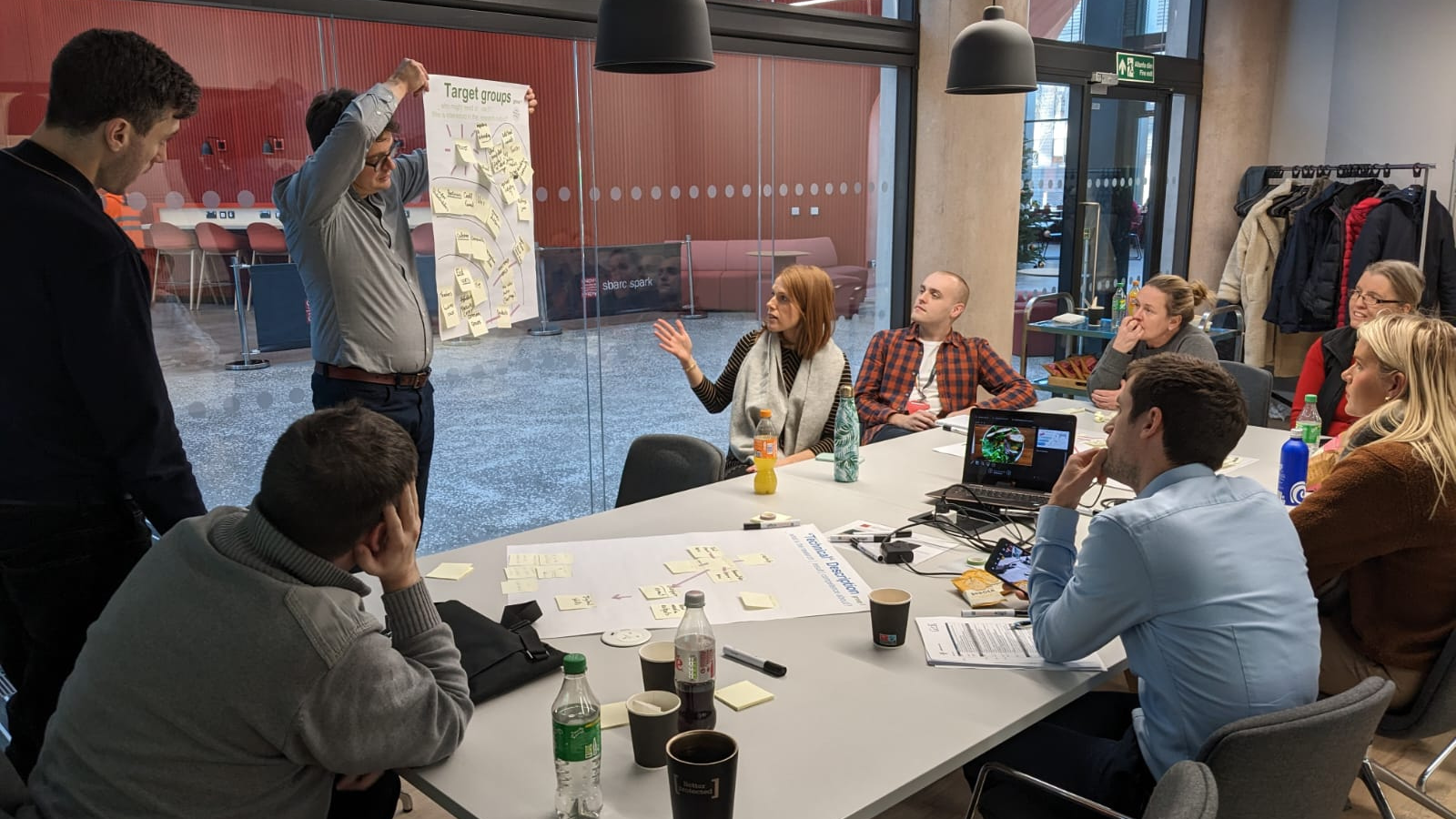 Group of people sat around a workshop table looking at a man holding up a poster
