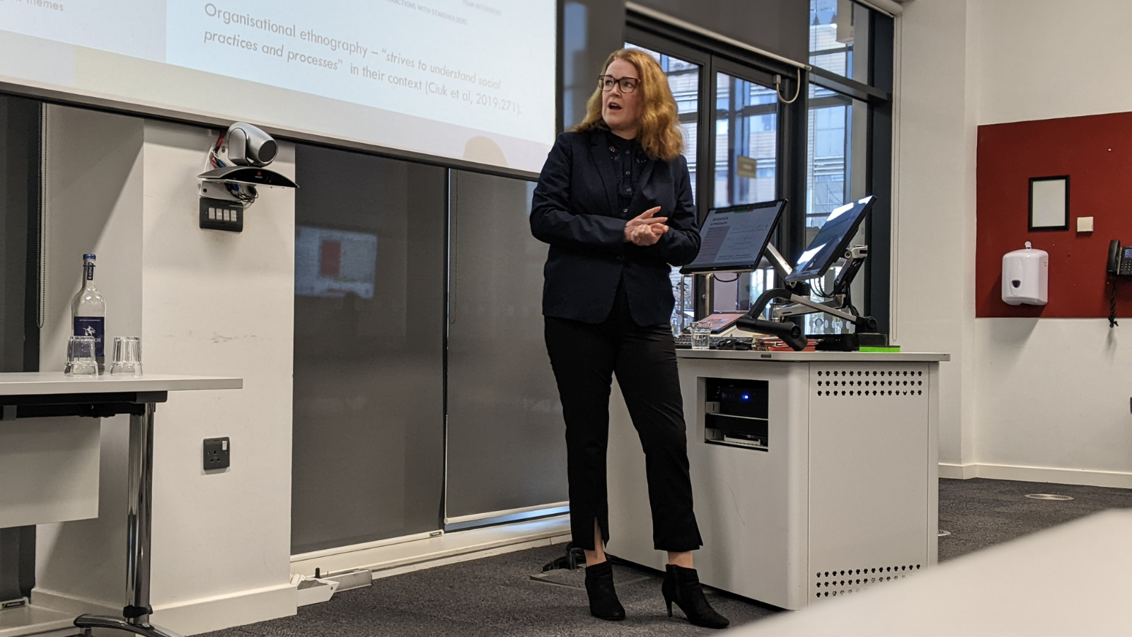 A lady stood in front of a large powerpoint screen presenting to an audience